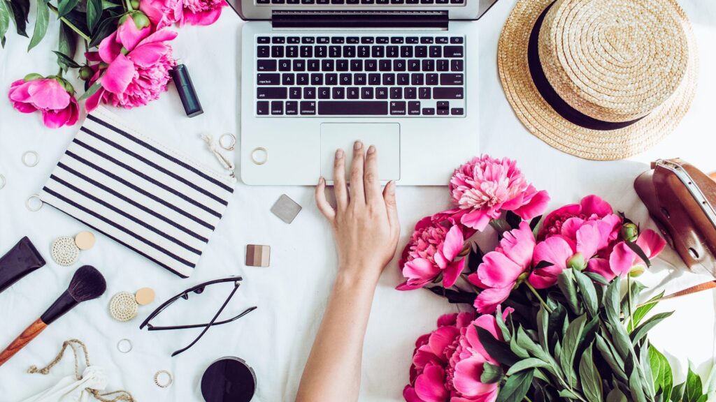 a woman's hand reaching for a laptop keyboard surrounded by makeup brushes, accessories, and a bouquet of flowers