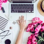 a woman's hand reaching for a laptop keyboard surrounded by makeup brushes, accessories, and a bouquet of flowers