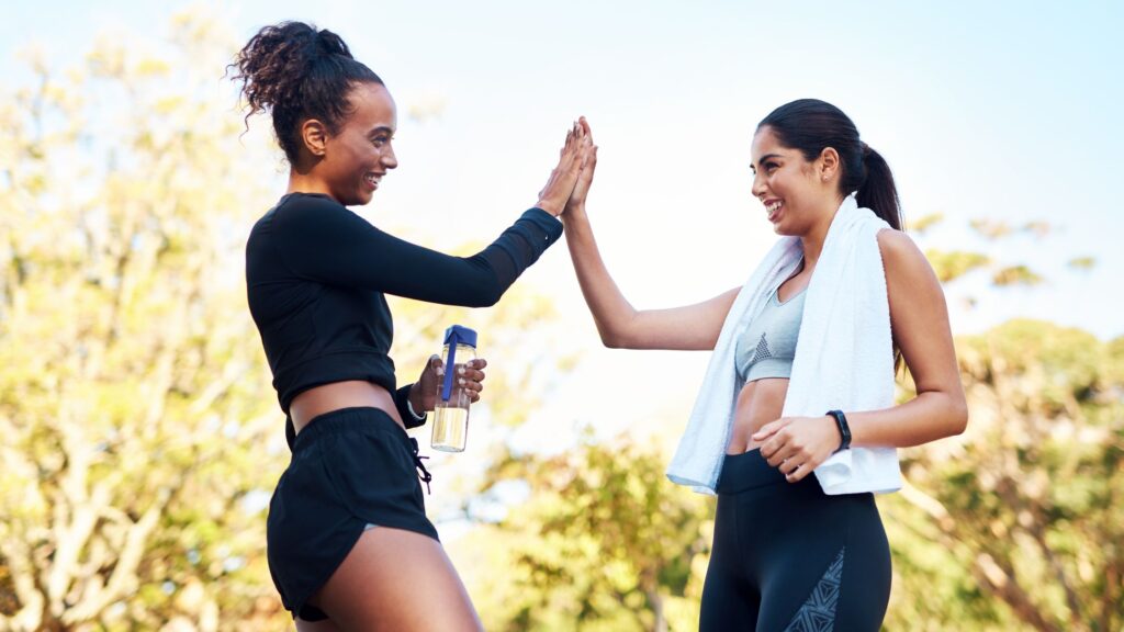 Two women in fitness clothes high-fiving in an outdoor backdrop.