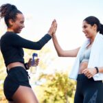 Two women in fitness clothes high-fiving in an outdoor backdrop.