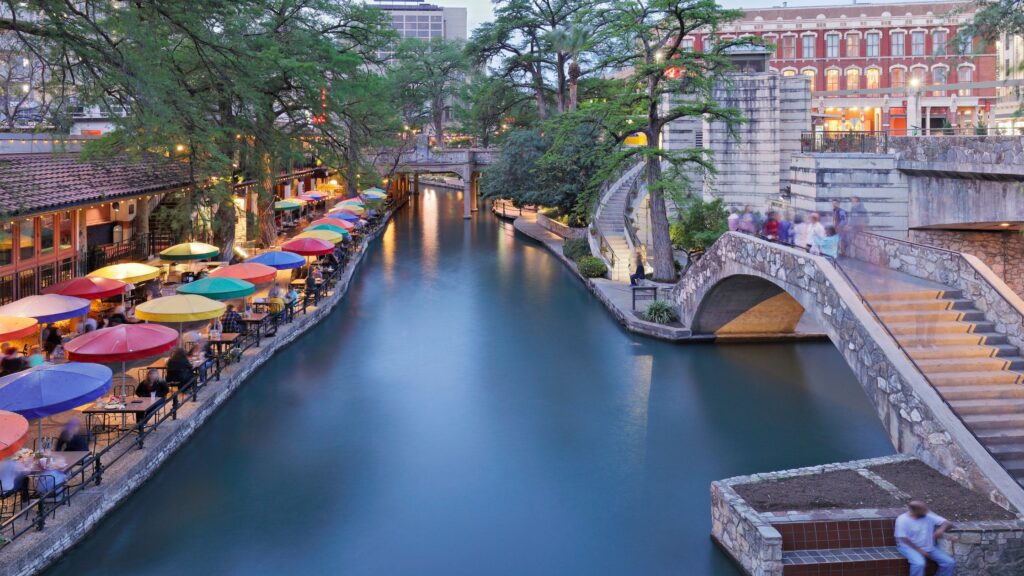 the riverwalk in san antonio, texas, lined with al fresco umbrella tables along the left bank.