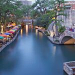 the riverwalk in san antonio, texas, lined with al fresco umbrella tables along the left bank.
