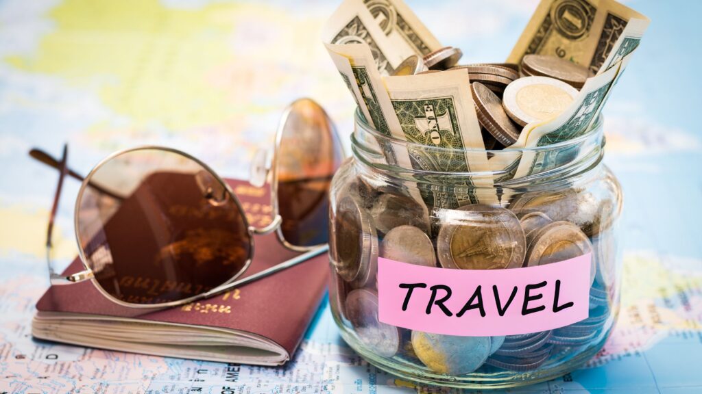 money jar with label “Travel” sitting on a U.S. Map next to a passport and pair of sunglasses