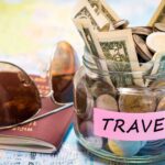 money jar with label “Travel” sitting on a U.S. Map next to a passport and pair of sunglasses