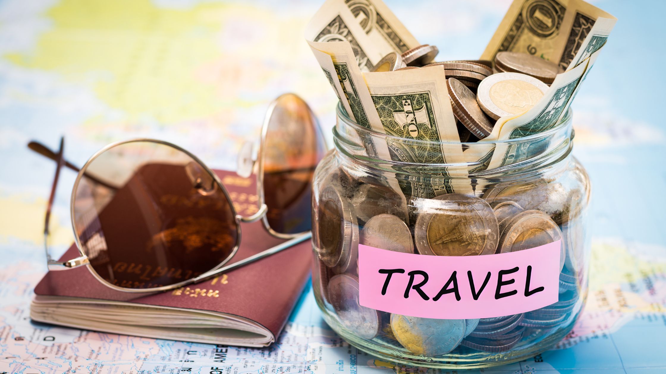 money jar with label “Travel” sitting on a U.S. Map next to a passport and pair of sunglasses