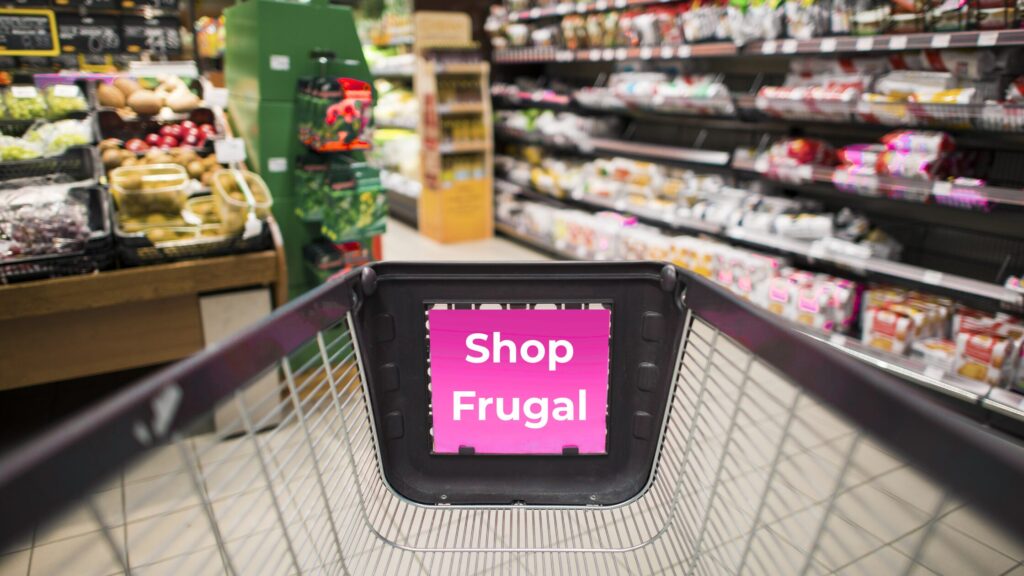 A close-up of an empty grocery cart being pushed through the produce section of a grocery store. The banner on the inside of the cart reads: Shop Frugal.