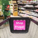 A close-up of an empty grocery cart being pushed through the produce section of a grocery store. The banner on the inside of the cart reads: Shop Frugal.
