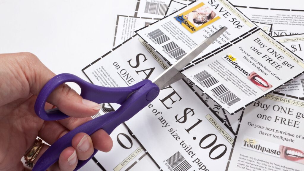 Hand holding purple scissors cutting paper grocery coupons on a white table