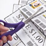 Hand holding purple scissors cutting paper grocery coupons on a white table