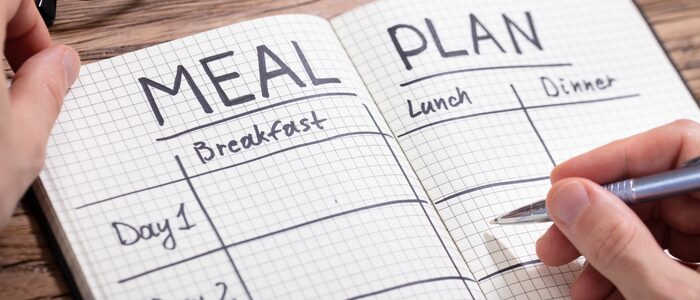 fh-inblog-meal-plan Meal planning notebook open with breakfast, lunch, and dinner sections written on graph paper while hands hold a pen over the page