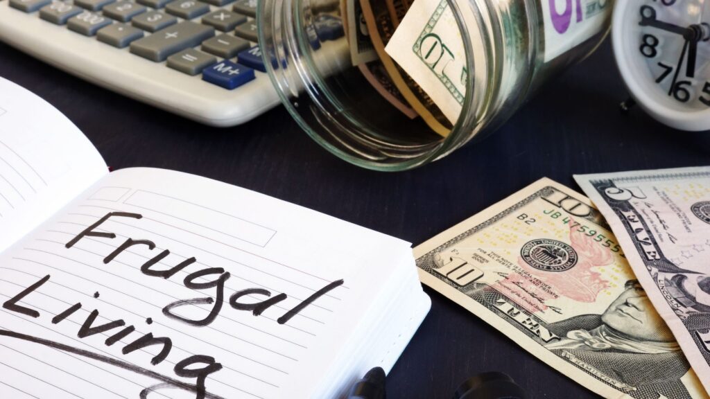 A notebook labeled “Frugal Living” beside a calculator, spilled jar of cash, and scattered dollar bills, symbolizing budgeting, saving, and mindful spending.