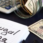 A notebook labeled “Frugal Living” beside a calculator, spilled jar of cash, and scattered dollar bills, symbolizing budgeting, saving, and mindful spending.