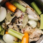 Overhead view of an Instant Pot filled with chicken bones, carrots, celery, onions, bay leaves, and peppercorns simmering in water to make homemade broth.