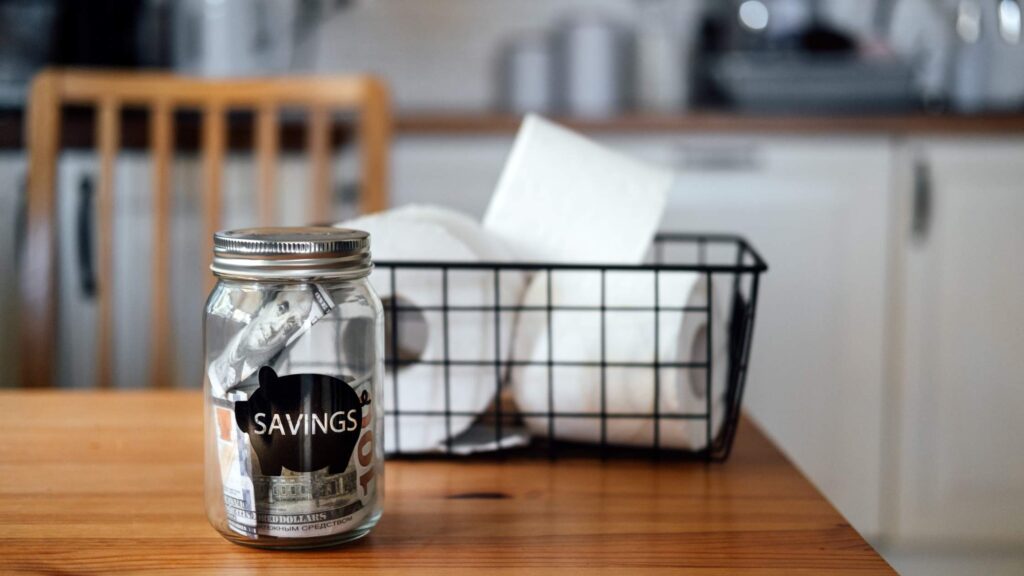 Savings jar with cash on a kitchen table next to a wire basket of toilet paper, showing practical habits for living frugally.