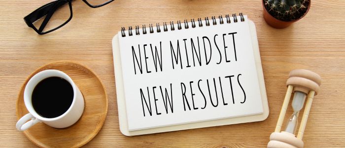 Notebook reading “New Mindset New Results” beside coffee cup and hourglass on desk, representing focus, growth, and the rewards of frugal living.