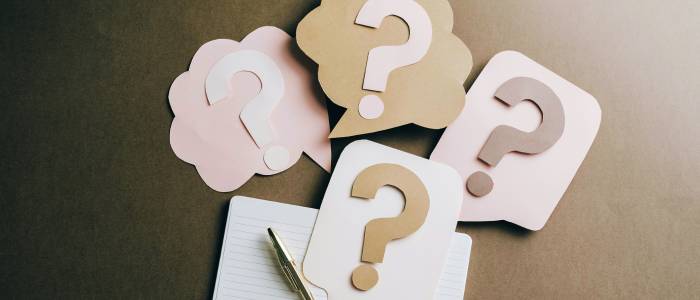 Paper cutout question marks in pastel speech bubbles arranged on a table above an open notebook with a pen.