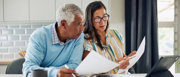 Older couple sitting at a table reviewing papers and discussing finances with a tablet nearby.