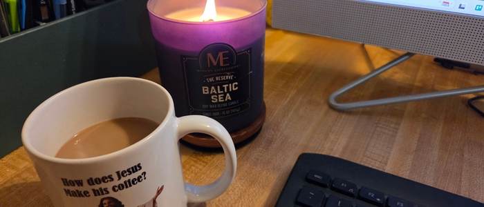 Coffee in a mug on a desk beside a lit candle and computer keyboard, representing a calm, frugal morning routine at home.