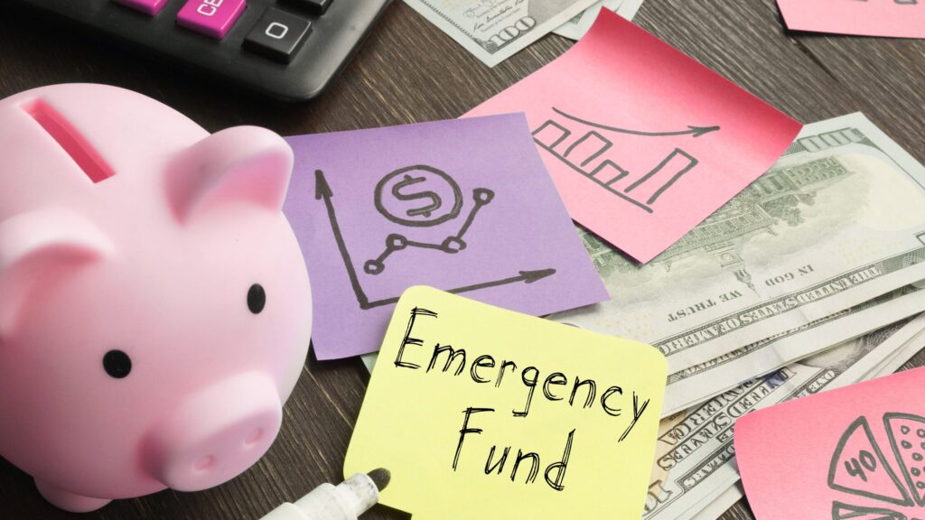 A small, pink piggy bank surrounded by a black keyboard with two pink keys, money, and multi-colored sticky notes with graphs. A yellow sticky note reads, Emergency Fund.