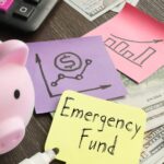 A small, pink piggy bank surrounded by a black keyboard with two pink keys, money, and multi-colored sticky notes with graphs. A yellow sticky note reads, Emergency Fund.