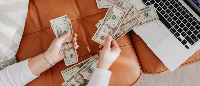 Close-up of a woman's hands while she counts money over leather cushions near an open laptop.