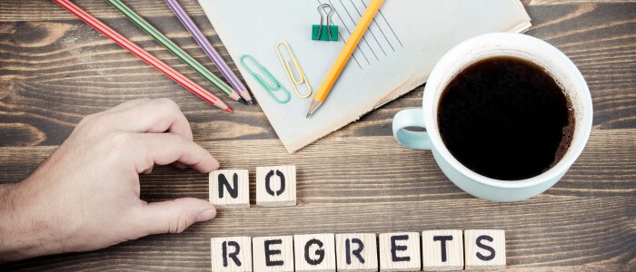 Hand arranging letter tiles spelling “NO REGRETS” on a wooden desk with coffee, notebook, and colored pencils nearby