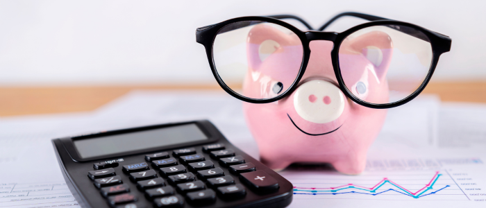 Pink piggy bank wearing black glasses beside a calculator on financial paperwork with small chart lines in the background.