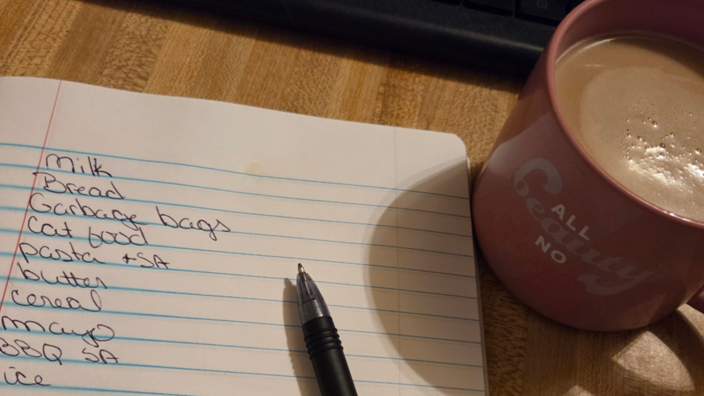 Handwritten grocery list beside a pink coffee mug and black pen on a wooden table.